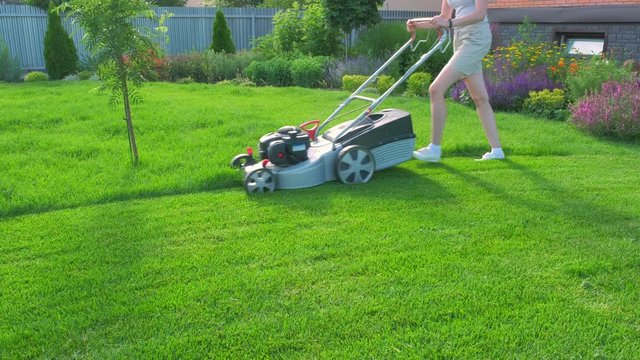 A woman mows green juicy grass with a lawn mower in the backyard of the house. Beautiful lush flower garden and young trees