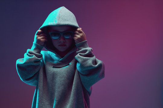 Portrait of a cool boy child in a rap image, stylishly posing in a hoodie, sunglasses and on a neon background.