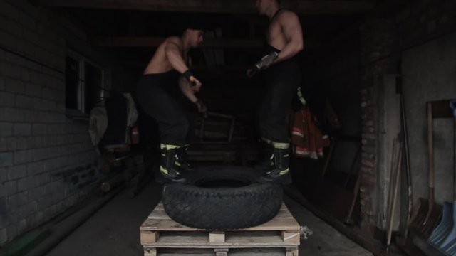 Two muscular young men are training and doing crossfit exercises by jumping to a high stance with a tire. The camera follows the movements of men