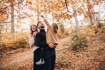 Stylish family in the autumn forest. Models. A young guy and a girl stand on a forest road among yellow leaves and hold their daughter in their arms.