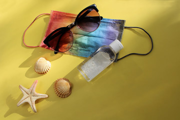Summer vacation fat lay with protective mask painted in rainbow colors, antiseptic, sunglasses, seashells and starfish on yellow background backlit by the sun. Travel and coronavirus covid-19 concept 