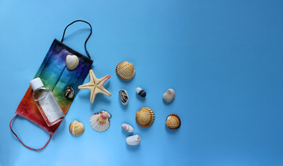 Summer vacation fat lay with protective mask painted in rainbow colors, antiseptic, seashells, pebbles and starfish on blue background. Travel and coronavirus covid-19 concept. Copy space