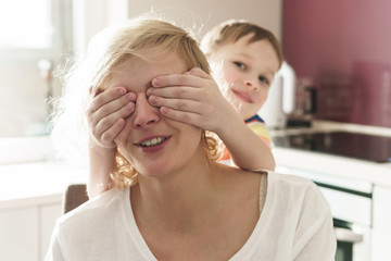 Cute boy is covering his mother's eyes on the kitchen