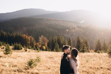 Stylish model couple in the autumn mountains. A young guy and a girl hug and kiss on a background of a forest and mountain peaks at sunset.