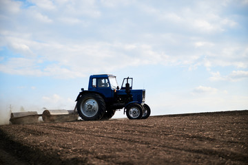 Obraz premium Agricultural work on field with black soil. Blue tractor riding and seeding countryside. Rural works in spring. Environmental protection concept. Ecological products cultivation. Natural background.