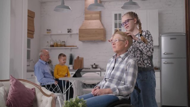 Close-up of handicapped elderly woman sitting in wheelchair while loving teenage granddaughter gently combing her hair. Cute little grandson with grandfather watching cartoons on background