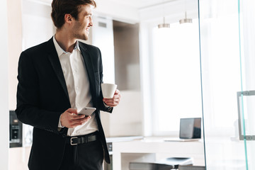 Photo of pleased businessman drinking coffee and using cellphone