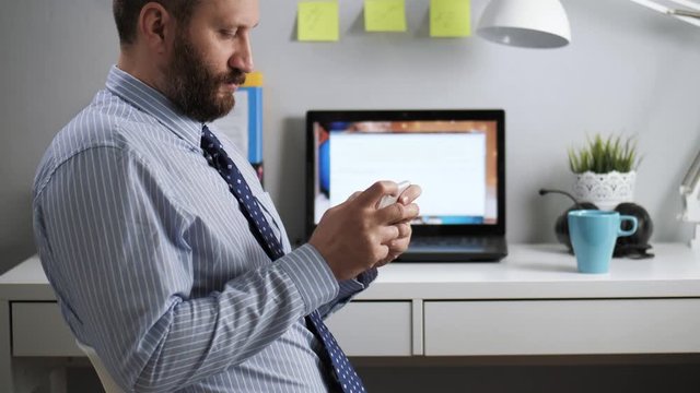 Office worker loafer is playing on phone. Bearded man sitting at workplace in office takes off from work and plays video games on mobile phone