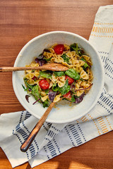 Top view of bowl with pasta salad and spatulas near napkin on wooden background