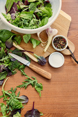 Top view of bowls with pepper, salt and salad ingredients on cutting board with knife on wooden background