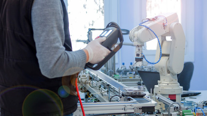Close-up robot arm, Man with Hand Gloves against COVID-19 ( Coronavirusis ) at background programming robot arm with teach pendant which is integrated on smart factory industry 4.0 manufacturing line.