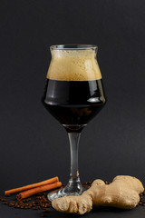 One tall glass of cold dark craft beer with spices over black background. Hops, anise, cinnamon, ginger. Copy space