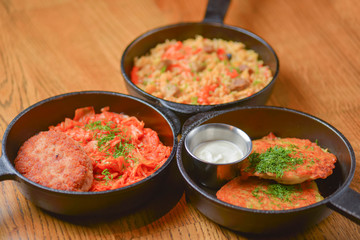 Delicious potato pancakes with sour cream, rice with meat and vegetables and Chicken Kiev cutlet served in frying pans.