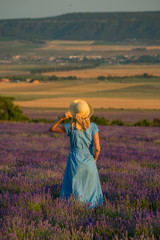 Slender young girl in a simple blue dress and a hat in a lavender field looks into the distance