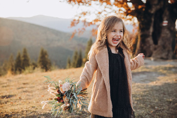 Portrait of a small stylish beautiful model girl who stands in the autumn mountains at sunset and holds a bouquet of flowers in her hands