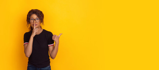 Finger on lips, pointing. African-american young woman's portrait isolated on yellow studio background. Beautiful female curly model. Concept of human emotions, facial expression, sales, ad, youth.