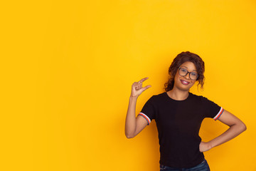 Shows something little, mockery. African-american young woman's portrait isolated on yellow studio background. Beautiful curly model. Concept of human emotions, facial expression, sales, ad, youth.