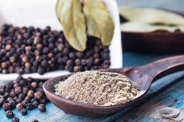 Ground black pepper in a spoon lies near the black peas scattered on the table and a bay leaf against the background of blue boards.