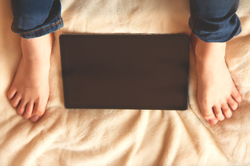Teenager sitting on the bed and looking at tablet screen. Tablet between legs. Home education. Selective focus. Top view