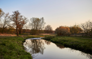 A curving stream with reflections, green meadow and an evening sky at sunset in spring in Brandenburg, Germany