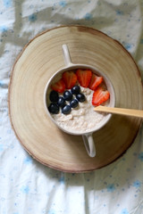 Bowl of vegan porridge with strawberry and blueberry topping, served in bed with floral sheets. Top view.