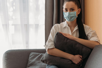 sad young woman in medical mask looking away while sitting on sofa and hugging pillow