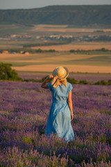 Slender young girl in a simple blue dress and a hat in a lavender field looks into the distance