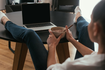 Obraz premium selective focus of girl holding bowl with breakfast while sitting with legs on table and looking at laptop with blank screen