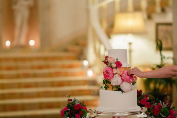 A wedding cake decorated with flowers. Woman makes final adjustments to bouquet, luxury hotel staircase in the background.  