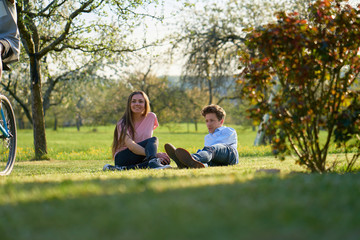 Young couple making a break in the country in Germany, laying on the ground on a meadow, looking after a cyclist, you can see trees and bushes