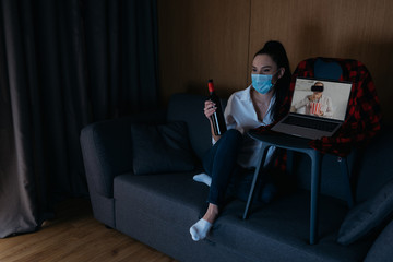 young woman in medical mask holding bottle of wine while sitting on sofa near laptop with boyfriend in vr headset on screen