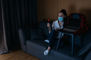 young woman in medical mask holding bottle of wine while sitting on sofa near laptop with blank screen