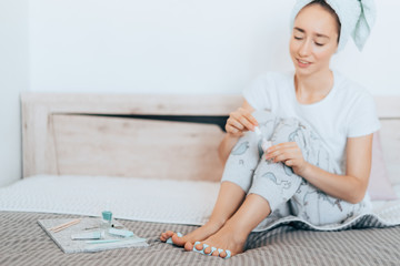 Girl sitting on the bed, making a pedicure herself and using a nail file after a shower at home because of the coronavirus epidemic. Home life during quarantine or self-isolation.