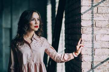 Fototapeta premium Beautiful young woman with curly dark hair and red lips in a powdery dress is standing near the window in the loft studio. Soft selective focus.