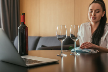 selective focus of woman sitting at table near wine glasses, bottle, medical mask and laptop
