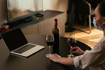 selective focus of young woman in medical mask near laptop with blank screen, glasses and bottle of red wine