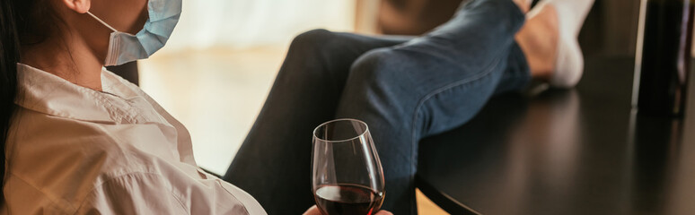 cropped view of woman in medical mask holding glass of red wine while sitting with legs on table, horizontal image