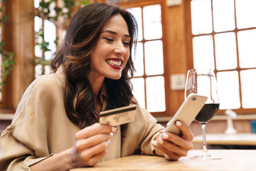 Image of excited adult woman holding credit card and using cellphone - obrazy, fototapety, plakaty