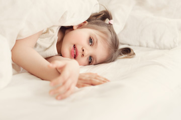 Obraz na płótnie Canvas Little cheerful girl 4 years old smiles and lies on bed on under white blanket, lifestyle