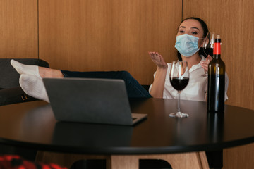 young woman in medical mask sending air kiss during video chat near bottle and glasses of red wine