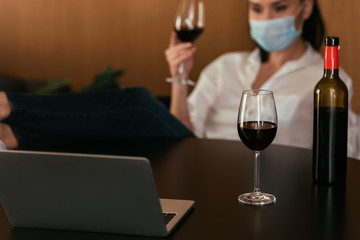 selective focus of woman in protective mask holding glass of red wine during video chat on laptop