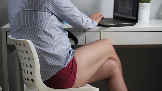 Remote work, freelance concept. Man in tie shirt headphones and underwear sitting at workplace in apartment surfing Internet on laptop