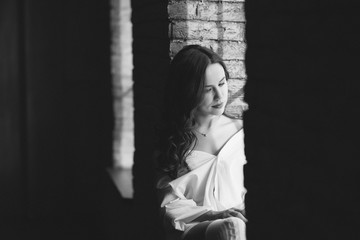 Fashionable young woman with perfect dark curly hair and beautiful make-up with puffy lips in an unbuttoned white shirt near the window. Soft selective focus. Black and white art photo.