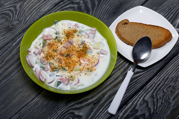 In a plate, chopped vegetables and kefir. Russian dish okroshka with kefir. Near a spoon and a piece of bread.