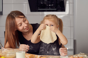 Mother and daughter preparing a sweet cake using flour, milk, sitting on chairs at a table in a...