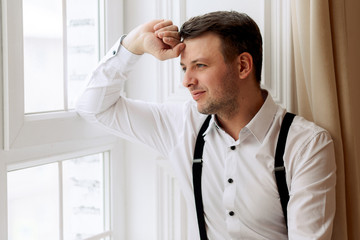 Fototapeta premium A young man of European appearance, by the window. Dressed in pants and a shirt. Portrait in a classic interior or studio. Businessman or freelancer. close-up, face.