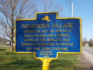 A New York state historic marker in dappled sunlight for Pathfinder Village in Edmeston, NY says it was the world's first community for residents with Down Syndrome