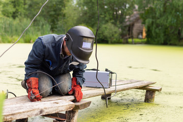 man welder in blue uniform works, welding mask and protective red gauntlets, metal welding with a welding machine, wooden bridge in the swamp.