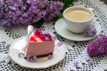 A berry mousse cake with chocolate base and juice jelly, decorated with lilac flowers, served with coffee.