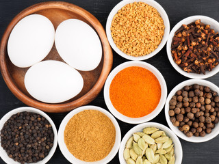 Raw chicken eggs, black pepper, masala powder, allspice, fenugreek, green cardamom, hot red chili pepper, cloves on black wooden background. Ingredients for making scrambled eggs or omelet
